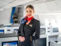 Uśmiechnięta stewardesa w mundurze LOT-u stoi na tle kabiny samolotu z rzędami foteli.