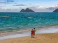 Para, gdzie na podróż poślubną. Para na plaży obserwuje łodzie z wioślarzami na turkusowym morzu, w tle góry.