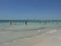 Słoneczny dzień na plaży Es Trenc. Ludzie kąpią się w turkusowej wodzie, a na horyzoncie widać żaglówki.