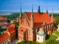 Gotycka ceglana katedra z zielonymi wieżyczkami i czerwoną dachówką, otoczona innymi budynkami i zielenią. Jedna z wielu pięknych katedr w Polsce.