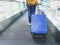 Osoba z niebieską walizką na ruchomych schodach lotniska. Zastanawiasz się, ile kosztuje odprawa na lotnisku?