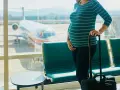 Czekając na lotnisku, przyszła mama zastanawia się, czy w ciąży można latać samolotem. Obok walizka, za oknem samolot.