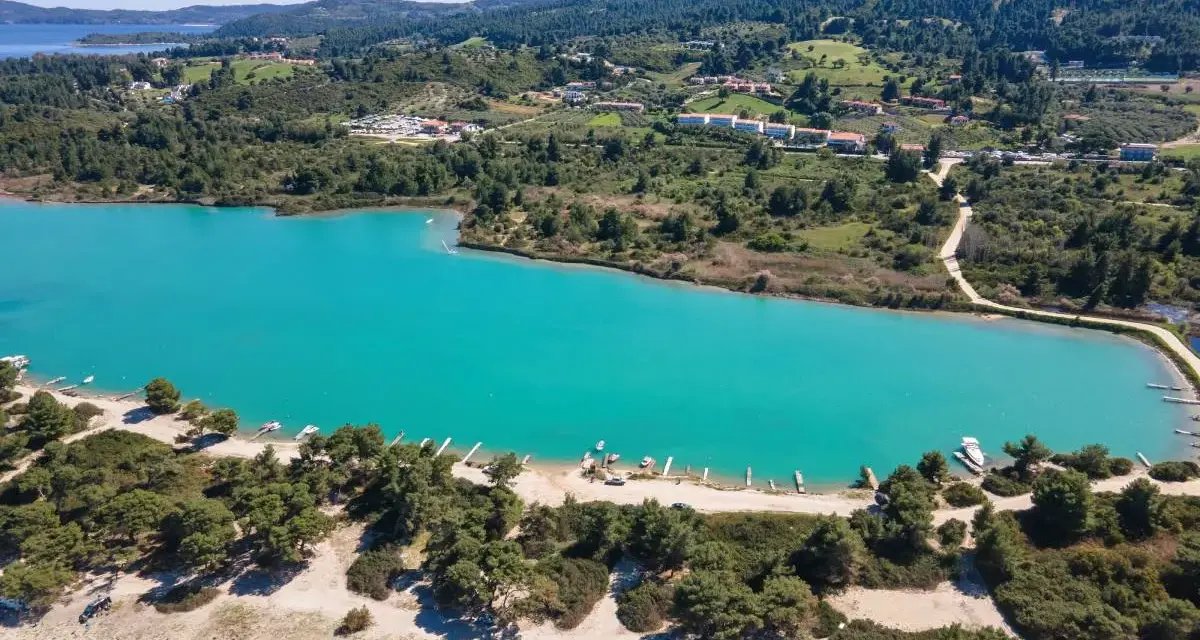 Turkusowa laguna na Kassandrze, otoczona zielonymi wzgórzami i lasem sosnowym. W oddali widać zabudowania i plażę z łodziami.