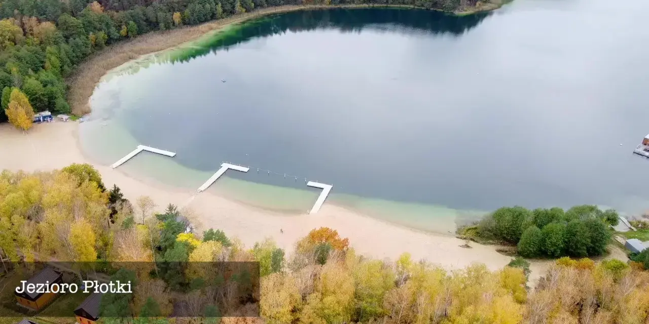 Jesioro Płotki jesienią. Piękna plaża, drewniane pomosty i kolorowe drzewa otaczają spokojną taflę wody.