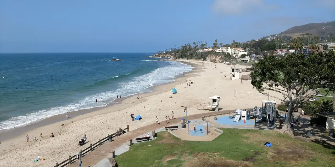 Słoneczny dzień na Laguna Beach. Ludzie spacerują po plaży i drewnianej promenadzie, dzieci bawią się na placu zabaw.