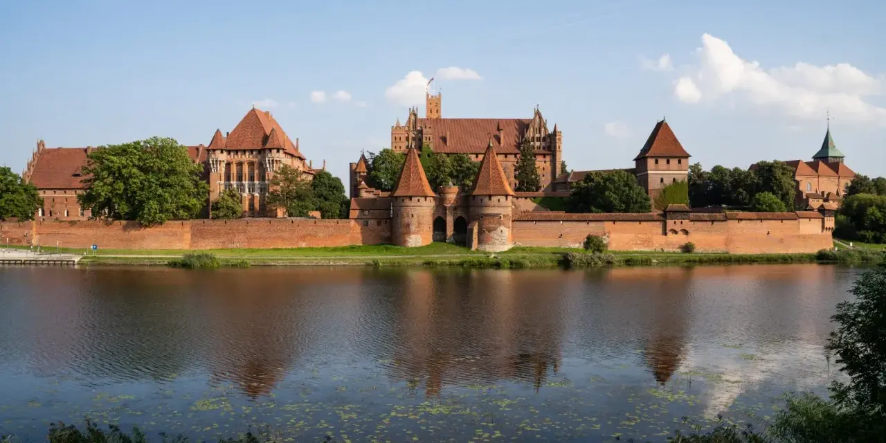 Potężny, ceglany zamek nad rzeką, największy zamek w Europie, z wieżami i murami obronnymi, odbija się w wodzie.