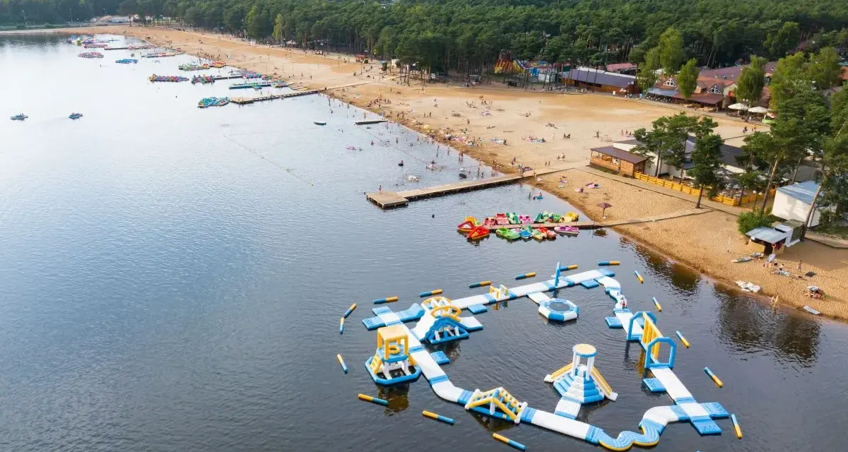 Sielpia jezioro tętni życiem. Na piaszczystej plaży wypoczywają ludzie, a w wodzie szaleją na dmuchanych zjeżdżalniach i torach przeszkód.