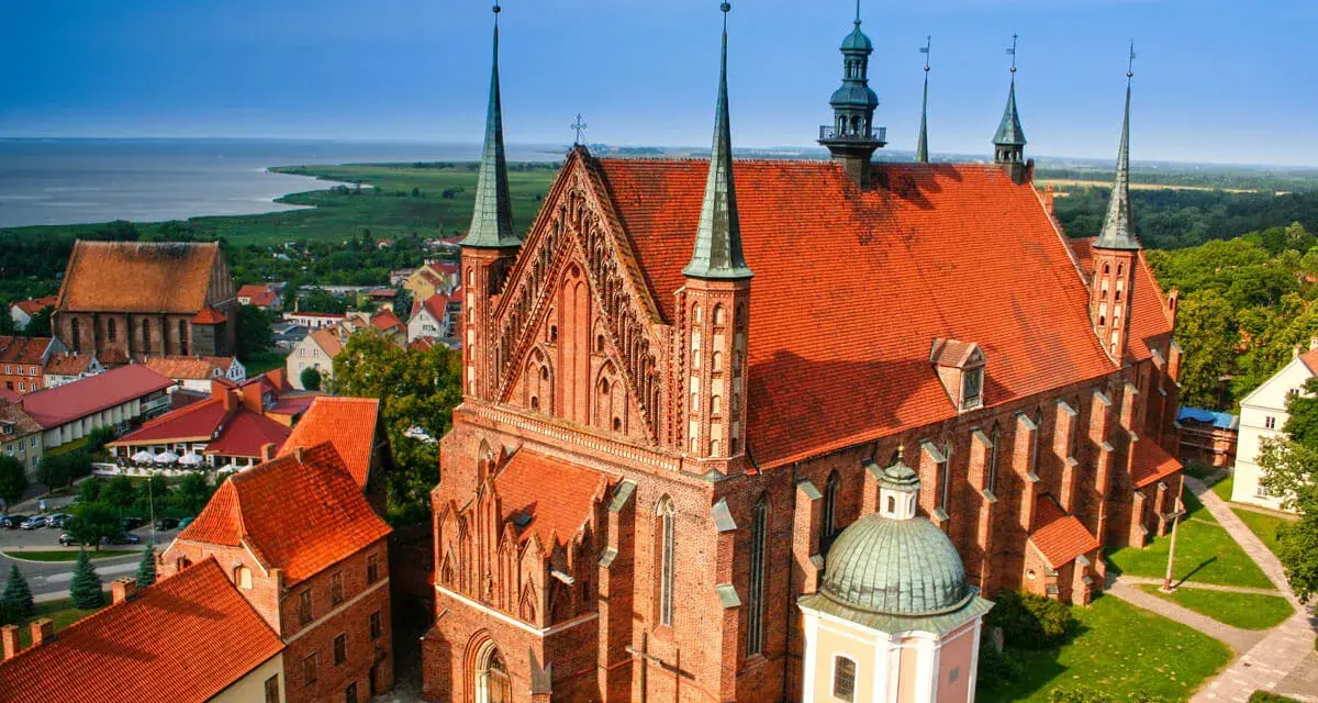 Gotycka ceglana katedra z zielonymi wieżyczkami i czerwoną dachówką, otoczona innymi budynkami i zielenią. Jedna z wielu pięknych katedr w Polsce.