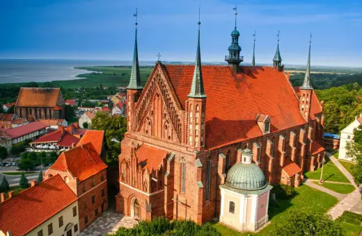 Gotycka ceglana katedra z zielonymi wieżyczkami i czerwoną dachówką, otoczona innymi budynkami i zielenią. Jedna z wielu pięknych katedr w Polsce.