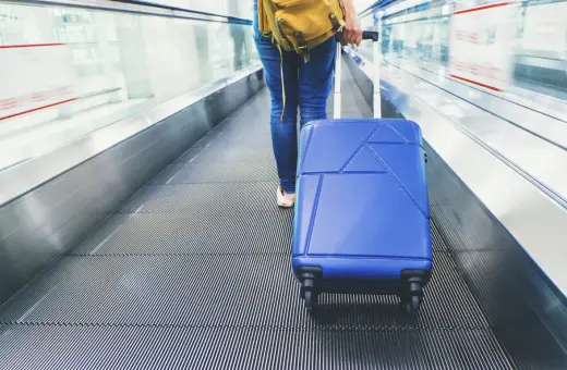 Osoba z niebieską walizką na ruchomych schodach lotniska. Zastanawiasz się, ile kosztuje odprawa na lotnisku?