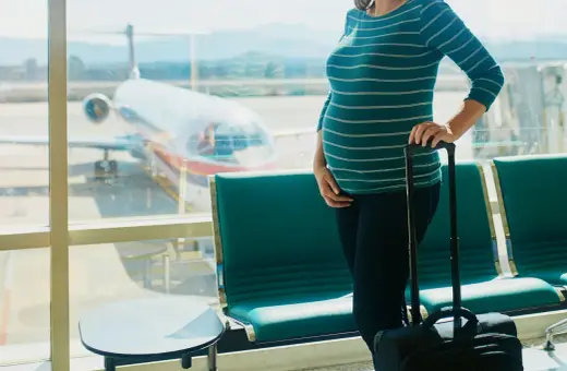Czekając na lotnisku, przyszła mama zastanawia się, czy w ciąży można latać samolotem. Obok walizka, za oknem samolot.
