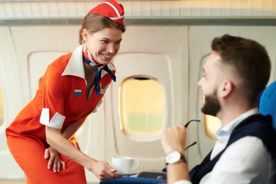 Uśmiechnięta stewardessa w pomarańczowym stroju podaje kawę pasażerowi. To jej praca, by podróż była komfortowa.