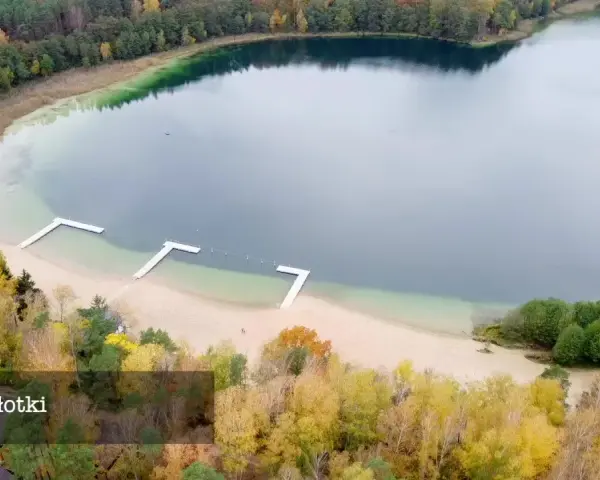 Jesioro Płotki jesienią. Piękna plaża, drewniane pomosty i kolorowe drzewa otaczają spokojną taflę wody.