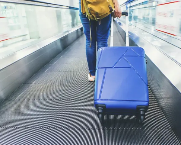 Osoba z niebieską walizką na ruchomych schodach lotniska. Zastanawiasz się, ile kosztuje odprawa na lotnisku?