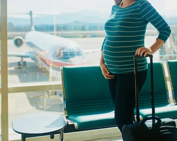Czekając na lotnisku, przyszła mama zastanawia się, czy w ciąży można latać samolotem. Obok walizka, za oknem samolot.