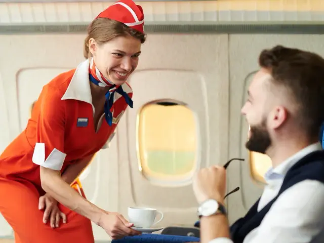 Uśmiechnięta stewardessa w pomarańczowym stroju podaje kawę pasażerowi. To jej praca, by podróż była komfortowa.