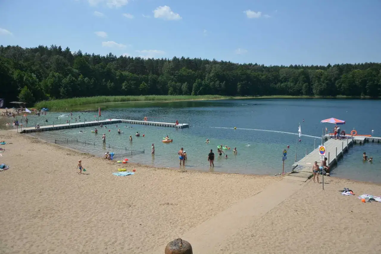 Słoneczny dzień nad jeziorem Płotki. Ludzie kąpią się, pływają na dmuchanych zabawkach i odpoczywają na piaszczystej plaży.