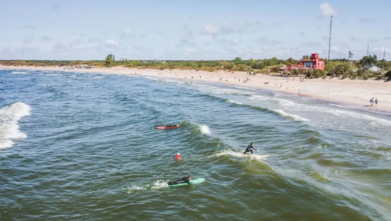 Surferzy na falach Bałtyku, słoneczny dzień na litewskim morzu. Plaża pełna ludzi, zielone wydmy i czerwony budynek w tle.