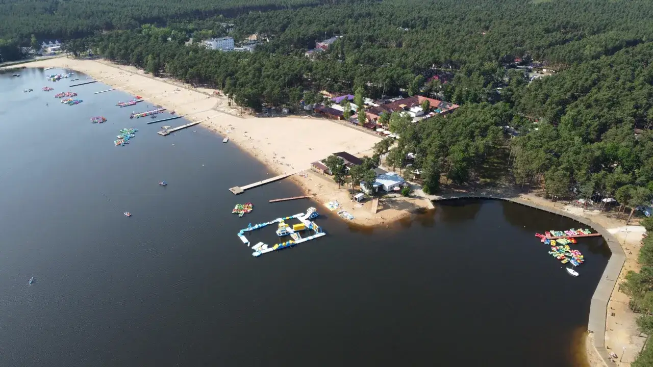 Sielpia jezioro tętni życiem. Na piaszczystej plaży wypoczywają ludzie, a w wodzie pływają kolorowe rowery wodne i dmuchane atrakcje.