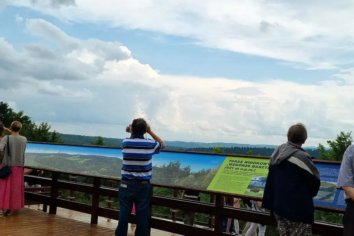 Ludzie podziwiają panoramę z tarasu widokowego na Wzgórzu Baszta. W oddali widać malownicze krajobrazy, a na pierwszym planie tablice informacyjne, w tym o kostrzynie.