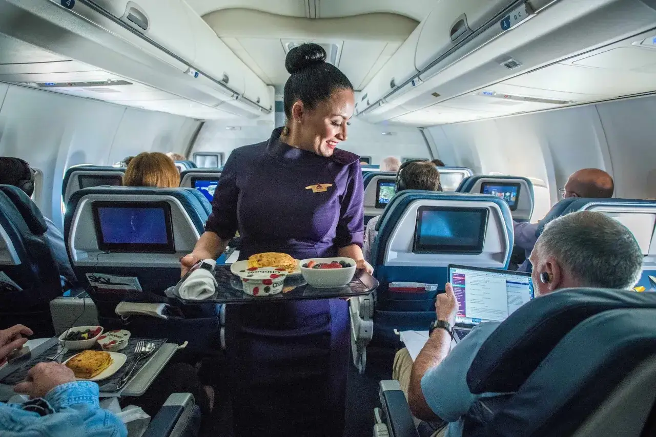 Uśmiechnięta członkini cabin crew serwuje posiłek w samolocie. Pasażerowie jedzą i pracują na laptopach.